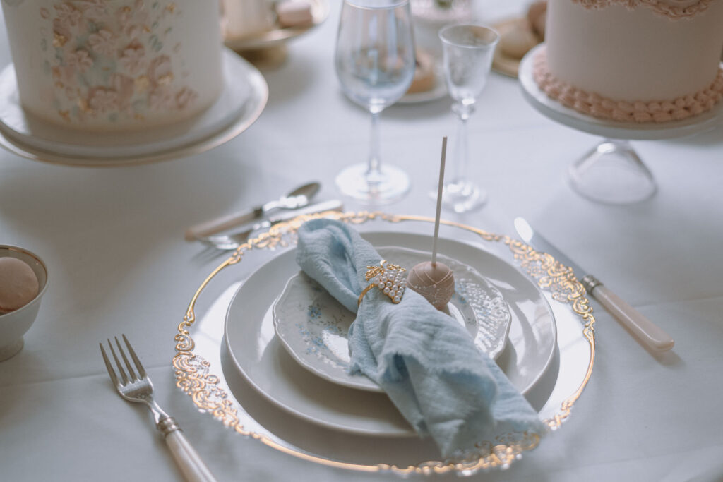 Elegant gedeckter Hochzeitstisch mit goldverziertem Teller, Stoffserviette, Cake Pop und edlen Details – Beispiel für hochwertige Gästebetreuung von Svenja Feddersen.