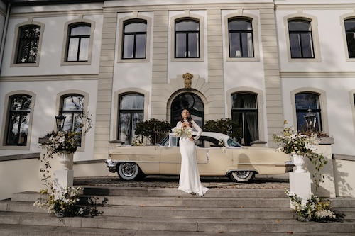 Braut vor einer eleganten Hochzeitslocation mit klassischem Gebäude und Vintage-Auto – Beispiel für exklusives Location Scouting von Svenja Feddersen.