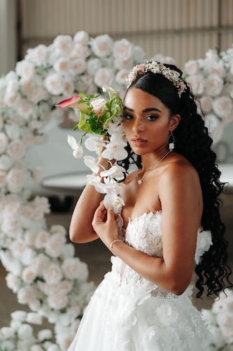 Braut in weißem Kleid posiert vor einem großen Blumenbogen und hält ein florales Arrangement.